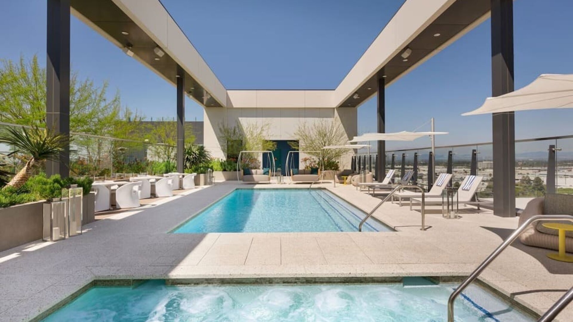 Rooftop pool deck featuring a swimming pool, spa area, sun loungers, shaded seating, and elevated views of the surrounding cityscape.